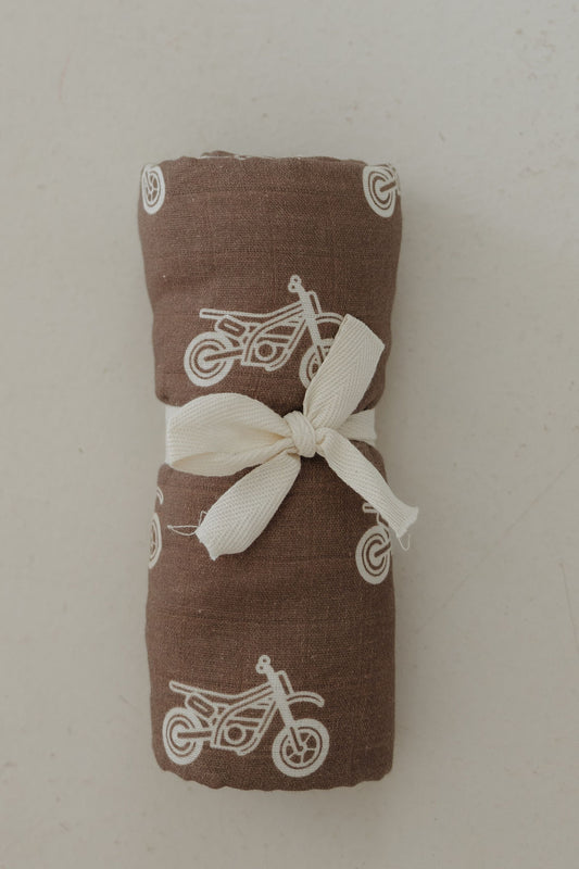 The forever french baby Muslin Swaddle | Dirt Bike features a brown muslin cotton fabric with white dirt bike and wheel prints, rolled neatly and tied with a cream ribbon, displayed on a light background.