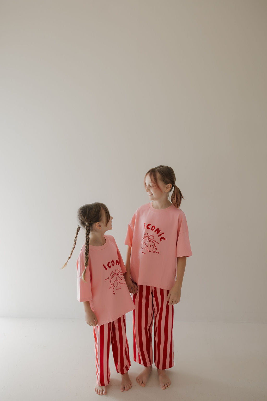 Two young girls smile indoors, holding hands in matching Minty Cherries T-Shirt & Pant Set | Iconic Cherry & Stripe—featuring a playful hand-drawn cherry design that’s perfect for any kids wardrobe.