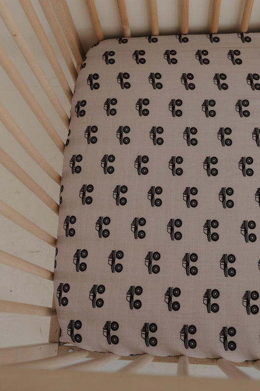 A close-up of a crib with light wooden slats featuring the forever french baby Muslin Crib Sheet | Monster Truck, decorated with a black and white monster truck pattern on a beige background.
