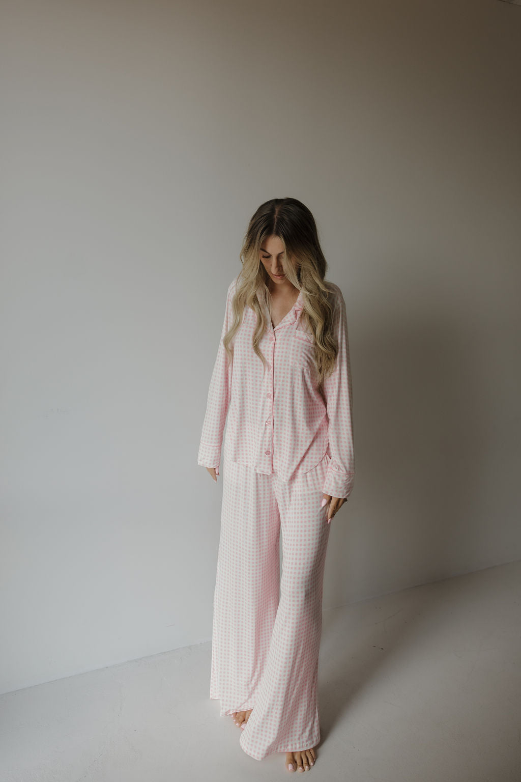 A woman with long, wavy hair stands barefoot indoors, wearing the forever french baby Women’s Bamboo Button Up Lounge Set | Giselle in a pink and white checkered pattern. She looks down as she touches her hair, enjoying the comfort and softness of this hypo-allergenic loungewear—perfect for relaxing at home.