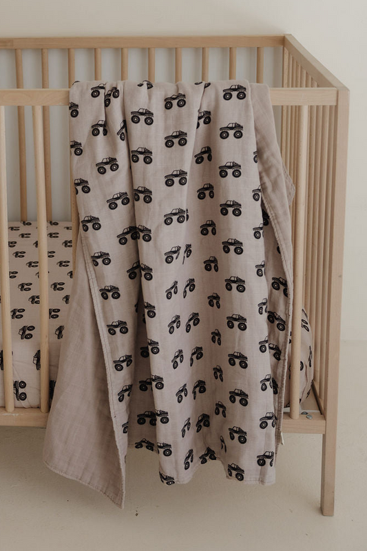 A light brown Muslin Quilt | Monster Truck by forever french baby, featuring a black monster truck pattern, hangs over a wooden crib with matching nursery bedding in a minimal, neutral setting.
