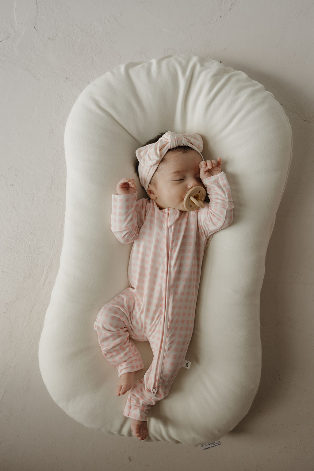 A baby in forever french baby’s Bamboo Zip Pajamas | Giselle, featuring a pink checkered pattern, sleeps peacefully on a white cushioned pad with a tan pacifier and matching headband. The hypoallergenic bamboo clothing adds to the serene, softly lit scene.