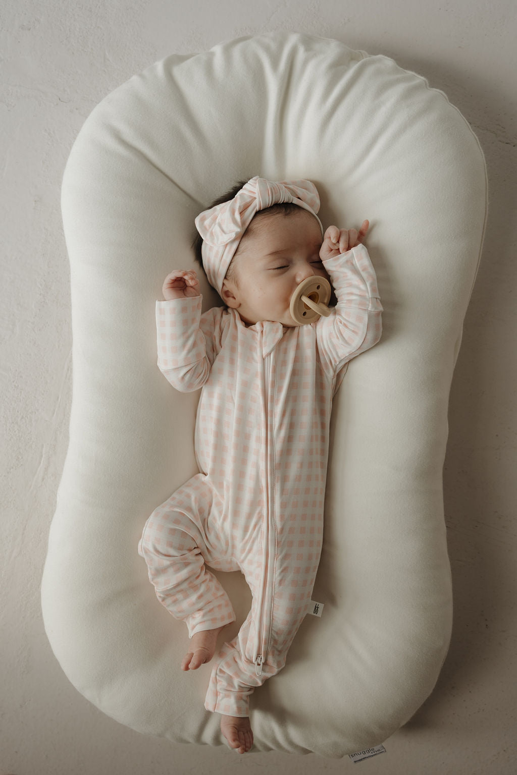 A baby in a pink checkered onesie and the Bamboo Head Wrap | Giselle from forever french baby sleeps on a white cushioned lounger, holding a tan pacifier in their mouth.