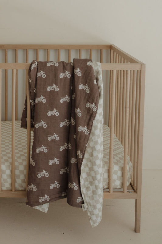 The forever french baby Muslin Quilt | Dirt Bike & Checkerboard, featuring a checkered sheet and a patterned brown blanket, is draped over a wooden crib and set against a plain, light wall.