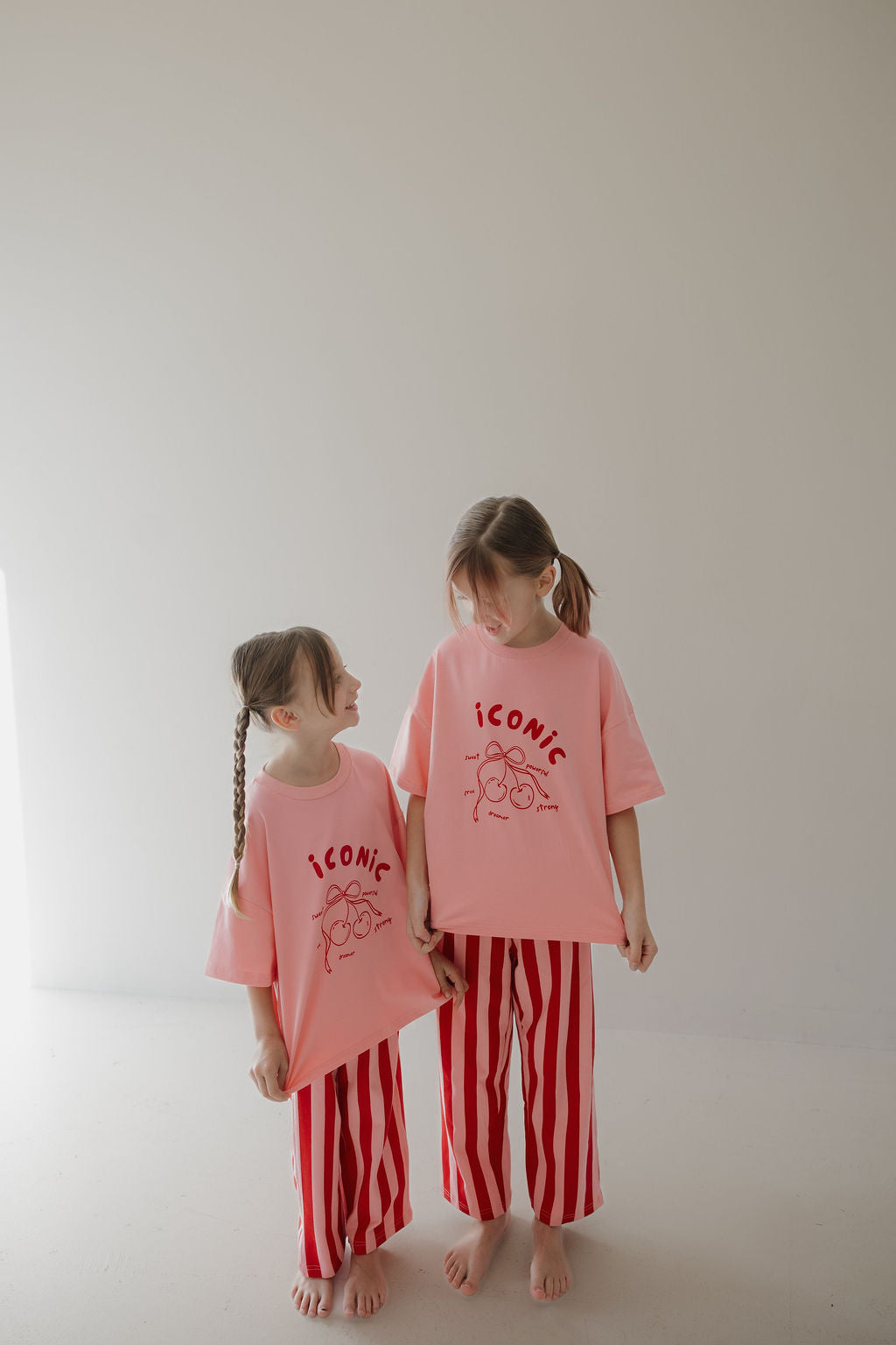Two girls with braided hair smile indoors in matching Minty Cherries T-Shirt & Pant Set | Iconic Cherry & Stripe, featuring a hand-drawn cherry motif and playful red-and-white stripes, against a plain background.