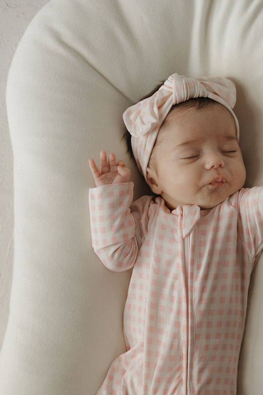 A baby wearing a pink and white checkered outfit and the Bamboo Head Wrap | Giselle from forever french baby sleeps peacefully on a soft white cushion, one arm raised beside their head.