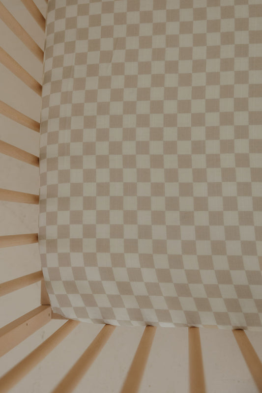 The Muslin Crib Sheet | Checkerboard by forever french baby features a soft beige and white checkerboard design, pairing perfectly with light wooden crib slats for cozy, stylish baby bedding.