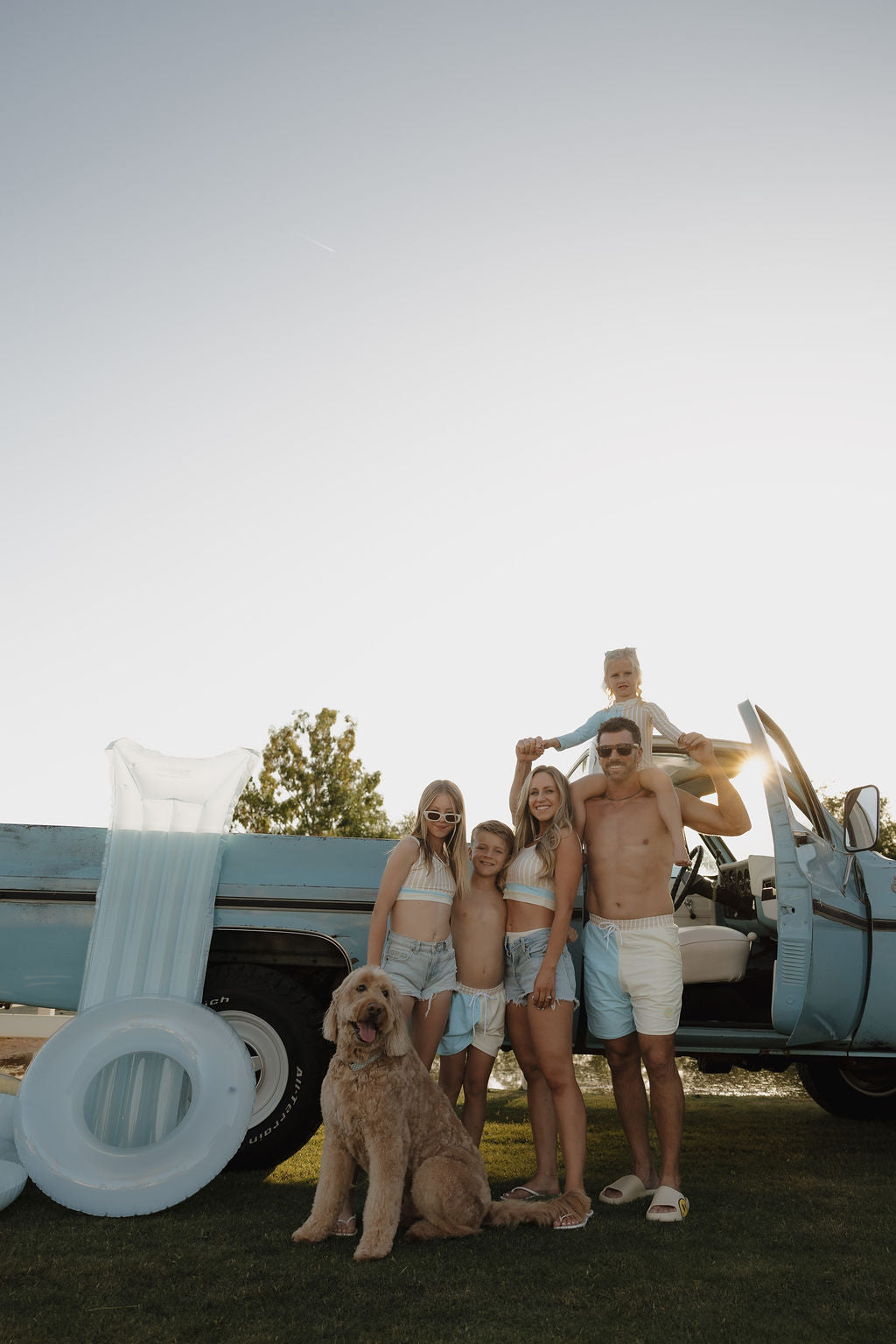 A family of five in summer wear poses by a vintage blue truck with pool floats, their large dog in front. Their daughter's lolo webb 2 Girl Long Sleeve Swimsuit | Surf & Sand radiates a warm, relaxed vibe at sunset.