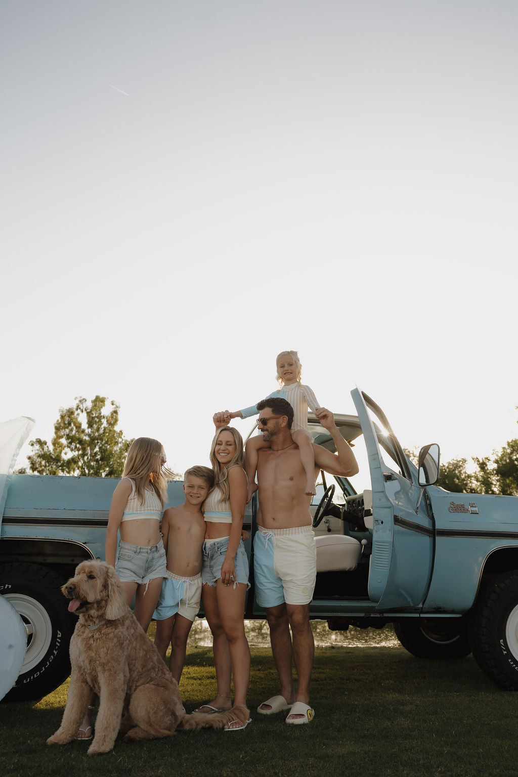 A family of five in matching lolo webb 2 Forever French outfits and a child wearing the lolo webb 2 Child Board Short | Surf & Sand smiles by a vintage blue truck, as their large dog sits proudly in front and dad lifts the youngest on his shoulders.