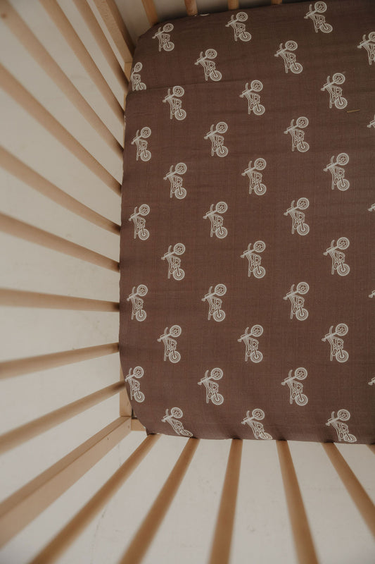 A crib with light wooden slats and nursery bedding, including the forever french baby Muslin Crib Sheet | Dirt Bike in brown with a white bear riding a bicycle line drawing pattern.