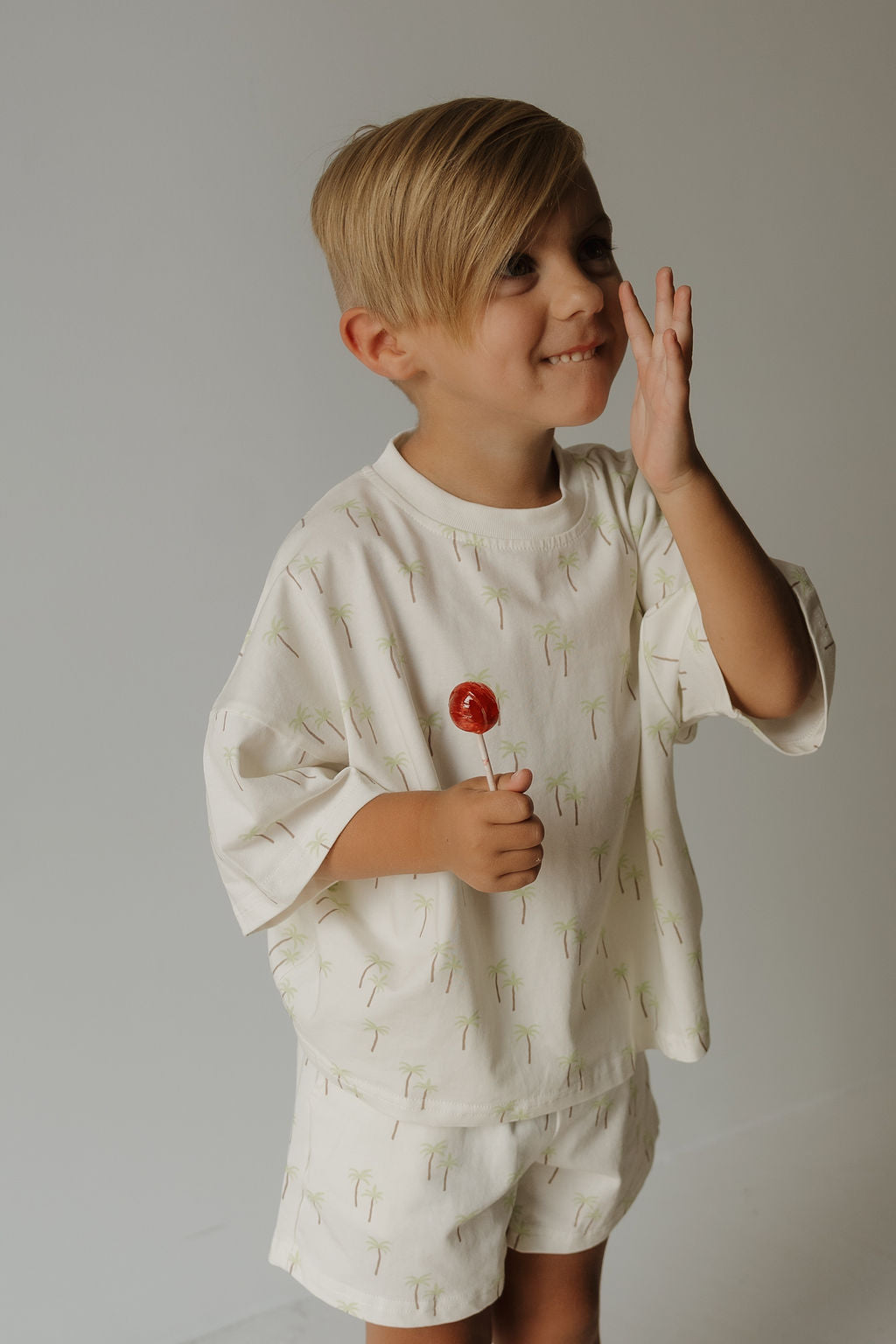 A young child with blond hair wears the forever french baby Child Short Set | Palm—comfortable playwear in a cream shade featuring a subtle palm tree print—as they smile, holding a red lollipop and touching their face with one hand against a plain background.