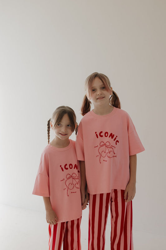 Two young girls with pigtails smile side by side in matching Minty Cherries T-Shirt & Pant Set | Iconic Cherry & Stripe, featuring "iconic" lettering on the tee and playful hand-drawn cherries on red-and-white striped pants.