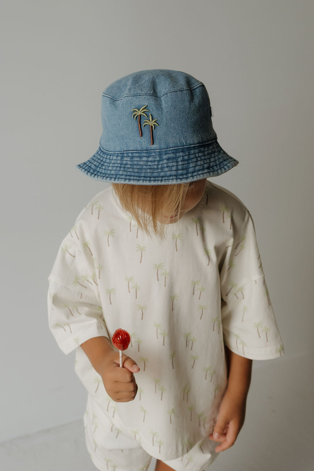 A young child wearing the forever french baby Bucket Hat | Palm, decorated with palm tree pins and paired with a coordinating palm tree print outfit, holds a red lollipop while looking slightly downward against a plain background.