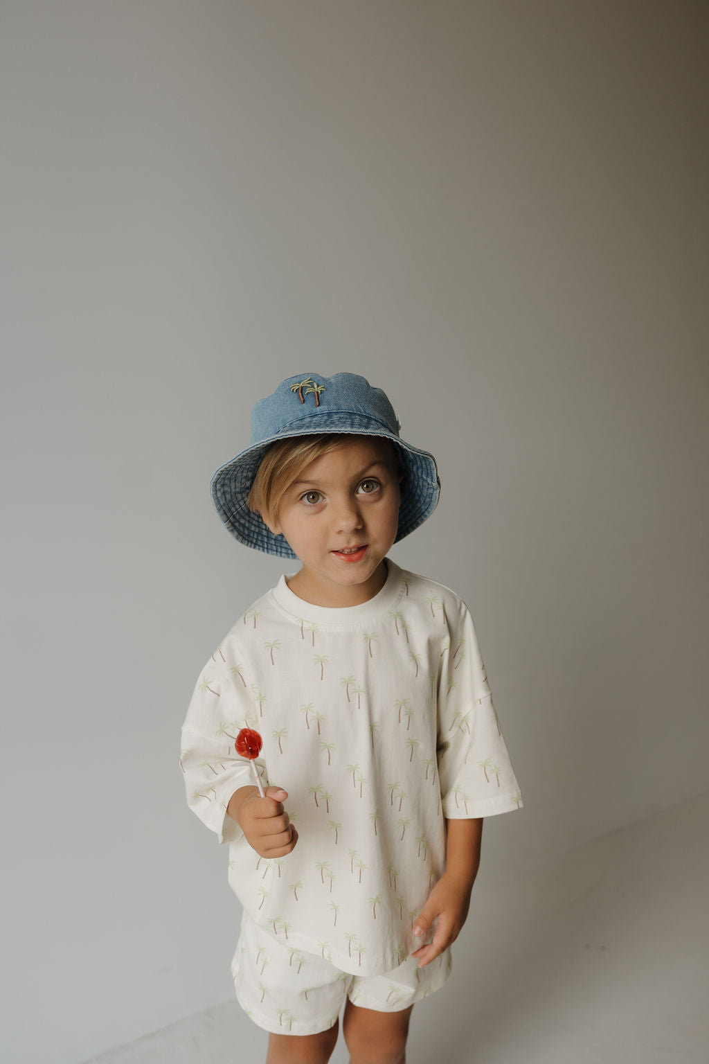 A young child wearing the Bucket Hat | Palm from forever french baby and a cream outfit with tiny designs stands against a plain background, holding a red lollipop and looking at the camera.