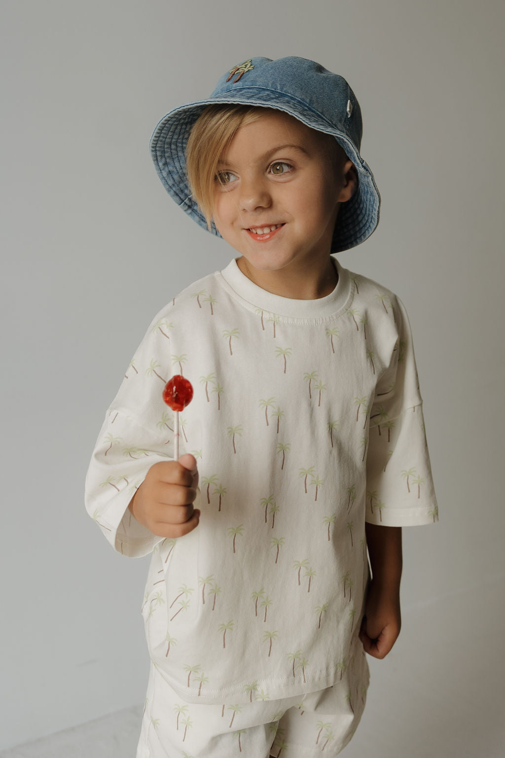 A smiling child wears the forever french baby Child Short Set | Palm—featuring a cream shirt with a palm tree print and matching playwear shorts—and a blue bucket hat, holding a red lollipop against a plain light background.