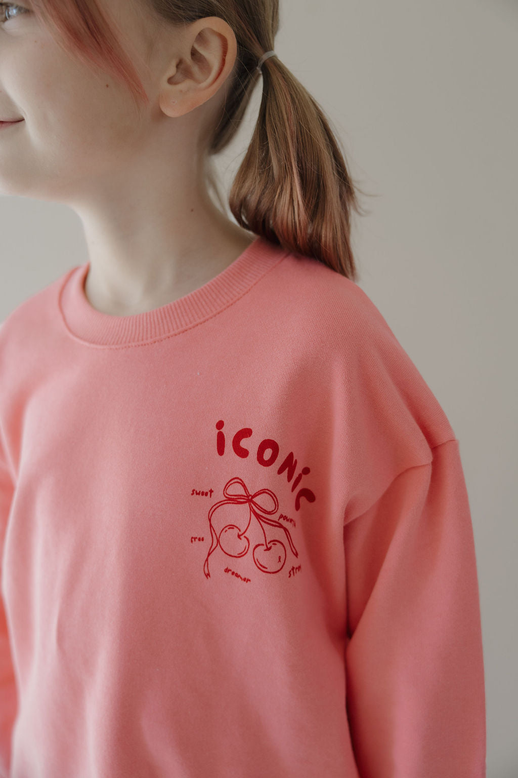 A smiling child with light brown hair in a ponytail wears the Minty Cherries Child Sweatshirt | Iconic Cherry, featuring a red hand-drawn cherry, "iconic," and handwritten words on the chest. The child is facing sideways.