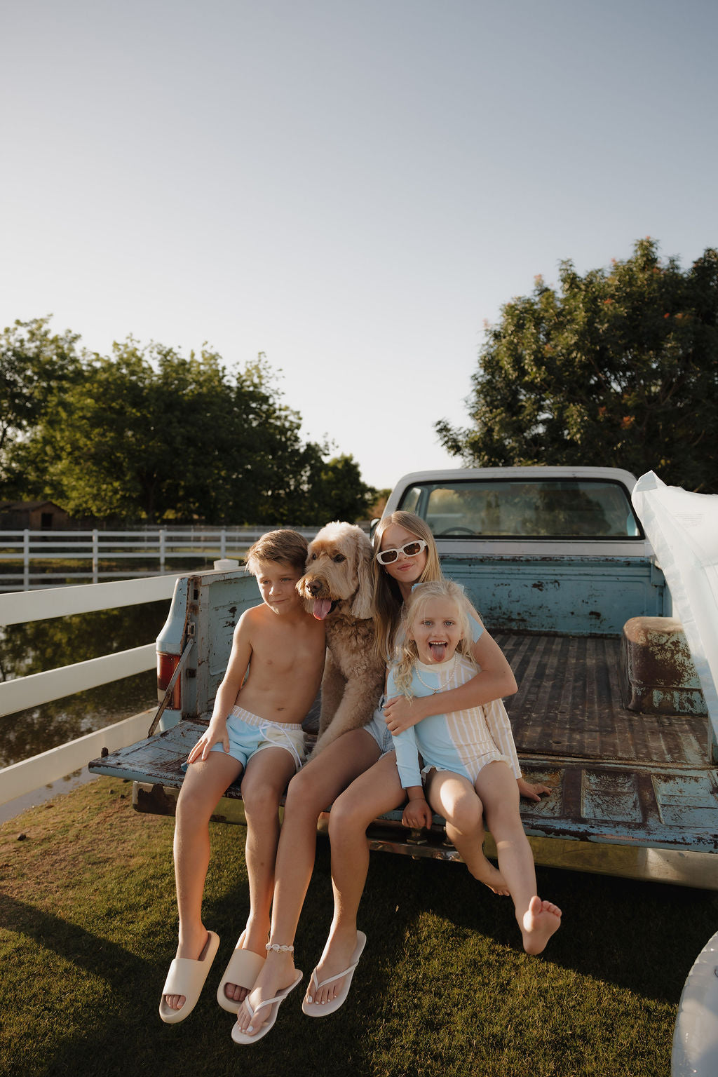 Three kids in swimsuits and lolo webb 2 Child Board Short | Surf & Sand sit on a blue truck tailgate with a fluffy dog, smiling and enjoying a sunny day outdoors near a white fence and trees.