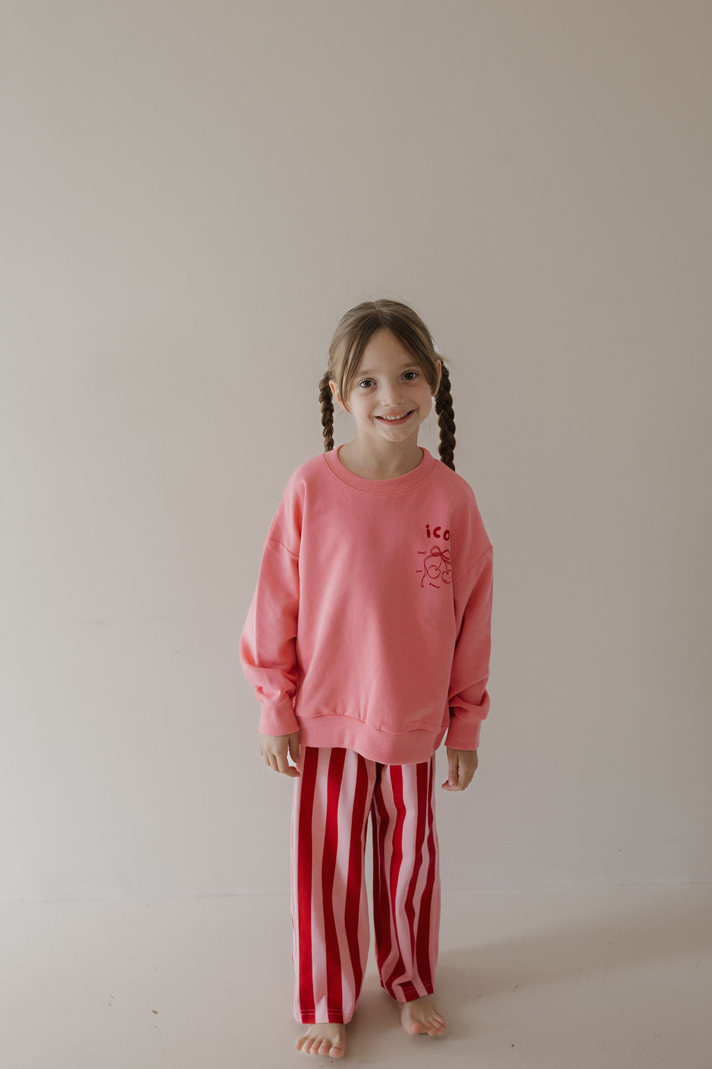 A young girl with brown hair in braids smiles barefoot against a plain background, wearing the Minty Cherries Child Sweatshirt | Iconic Cherry from the Miss Minty collaboration and red-and-white striped pants.