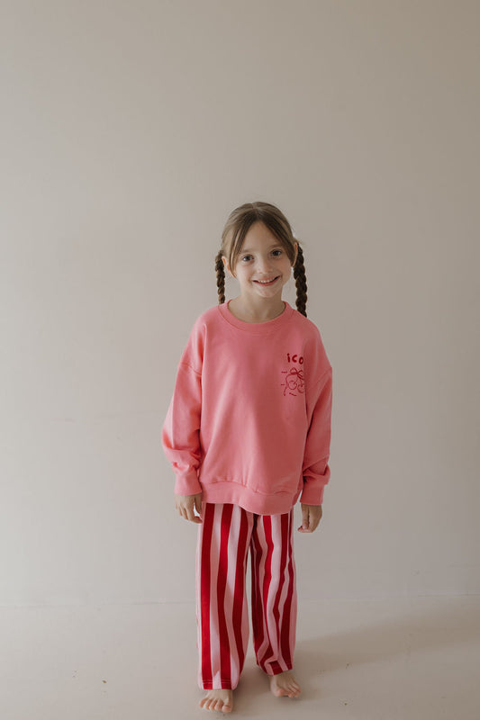 A young girl with brown hair in braids smiles barefoot against a plain background, wearing the Minty Cherries Child Sweatshirt | Iconic Cherry from the Miss Minty collaboration and red-and-white striped pants.