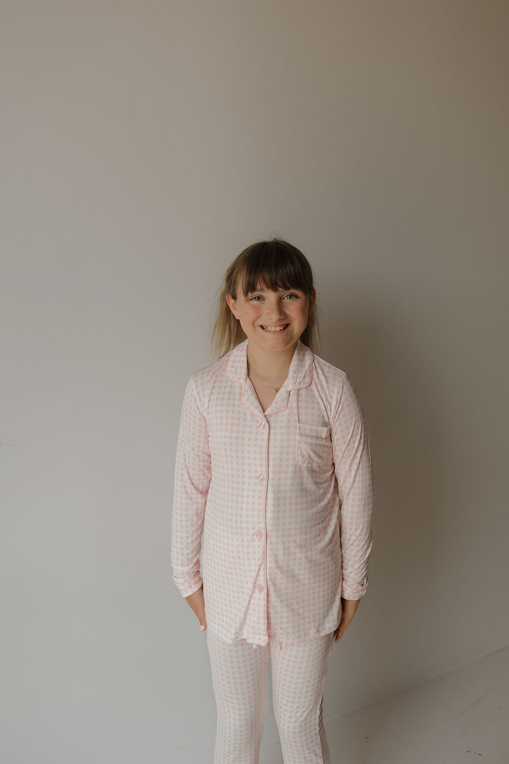 A young girl with light brown hair stands and smiles in front of a plain light-colored wall, enjoying the comfort of the soft, hypo-allergenic Girl’s Bamboo Button Up Lounge Set | Giselle from forever french baby.