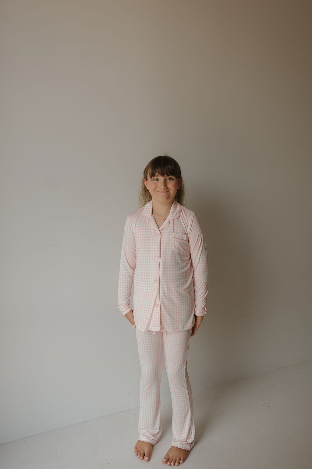 A young child stands barefoot against a plain wall, wearing the forever french baby Girl’s Bamboo Button Up Lounge Set in light pink and white check. The hypo-allergenic bamboo fabric is gentle on her skin, while her straight brown hair with bangs frames a soft smile for the camera.