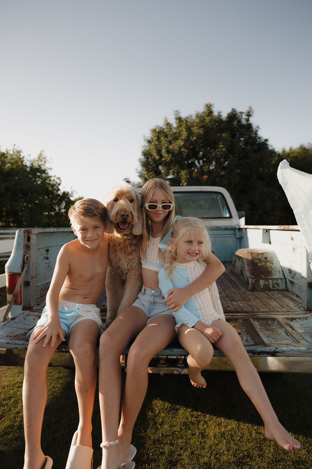 Three kids and a fluffy dog sit on a rusty pickup truck’s tailgate, smiling in the sun. They’re all wearing lolo webb 2 Child Board Short | Surf & Sand from the limited edition collection.