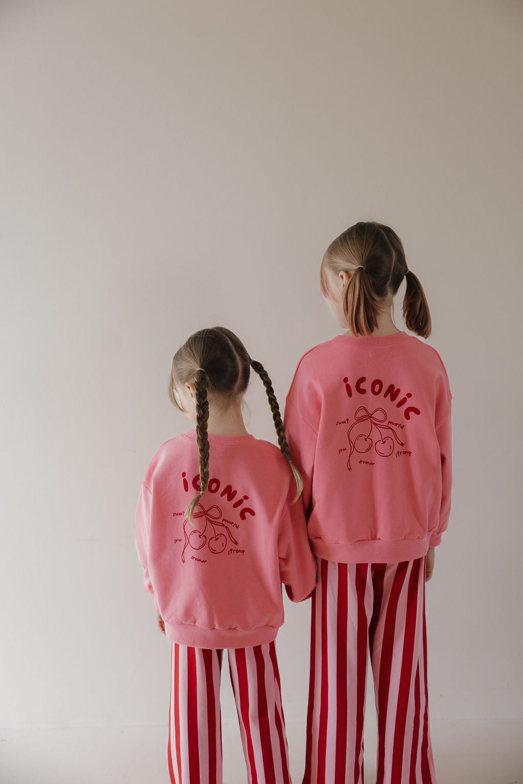 Two girls in matching Minty Cherries "Child Sweatshirt | Iconic Cherry" tops with hand-drawn cherry designs stand side by side, facing a beige wall. Their sweatshirts and red striped pants highlight the Miss Minty collaboration.