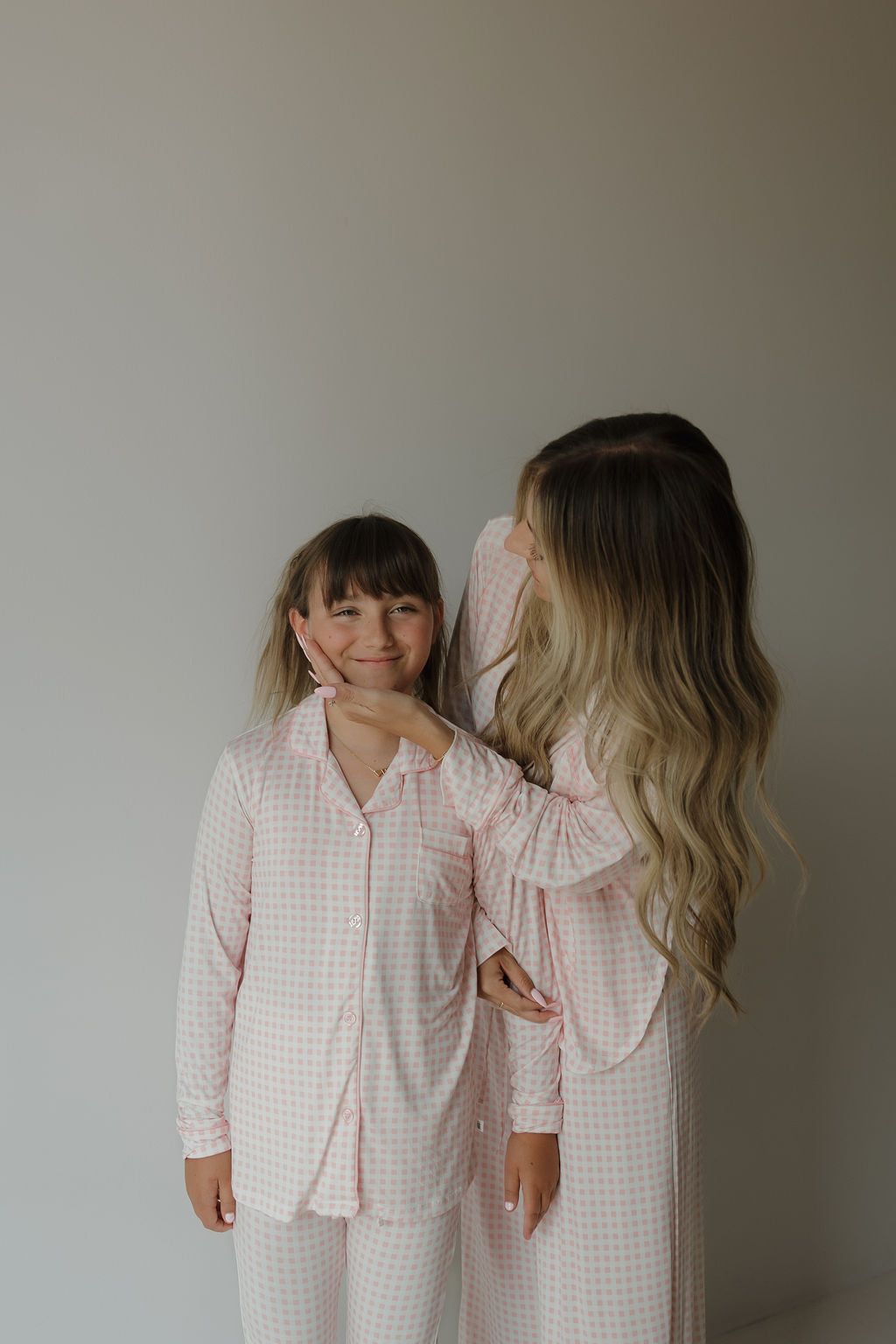 A woman with long blonde hair gently holds a smiling young girl’s chin as they stand against a plain light-colored background. Both are wearing matching hypo-allergenic bamboo pajamas from forever french baby, with the girl dressed in the Girl’s Bamboo Button Up Lounge Set | Giselle featuring a light pink and white checkered pattern.