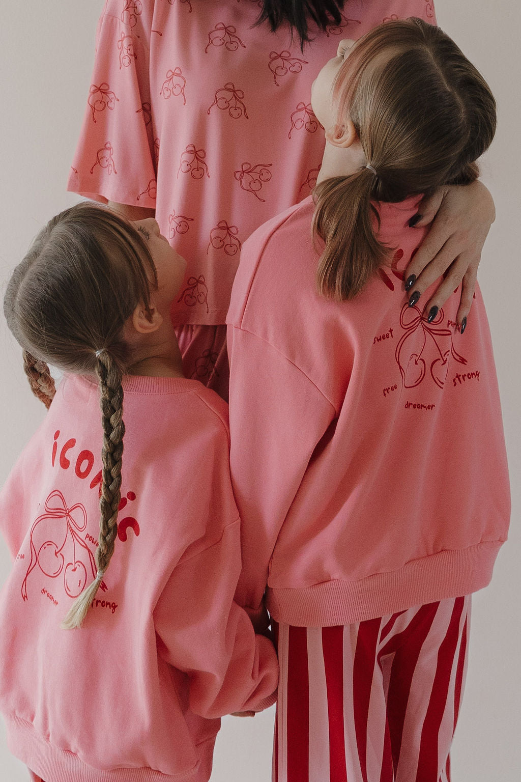 An adult and two children wear matching pink outfits; the kids sport Minty Cherries' Child Sweatshirt | Iconic Cherry, featuring a hand-drawn cherry design. The braided children hold hands, looking up at the adult in striped pink pants.