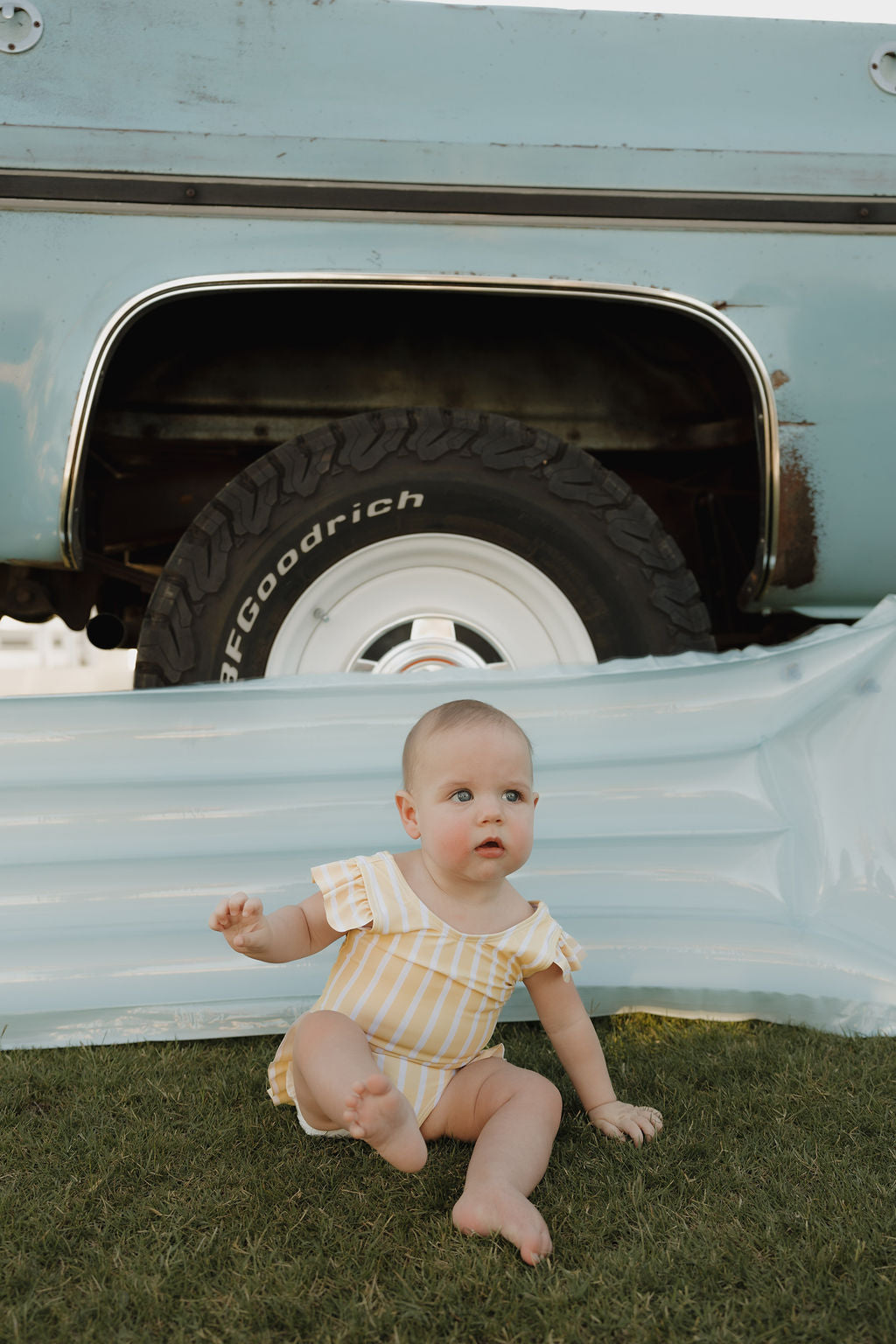 A baby in the lolo webb 2 Girl Sleeveless Swimsuit | Sun Rays sits on grass by a blue pool float and vintage truck tire, glancing curiously to the side—a charming summer moment.
