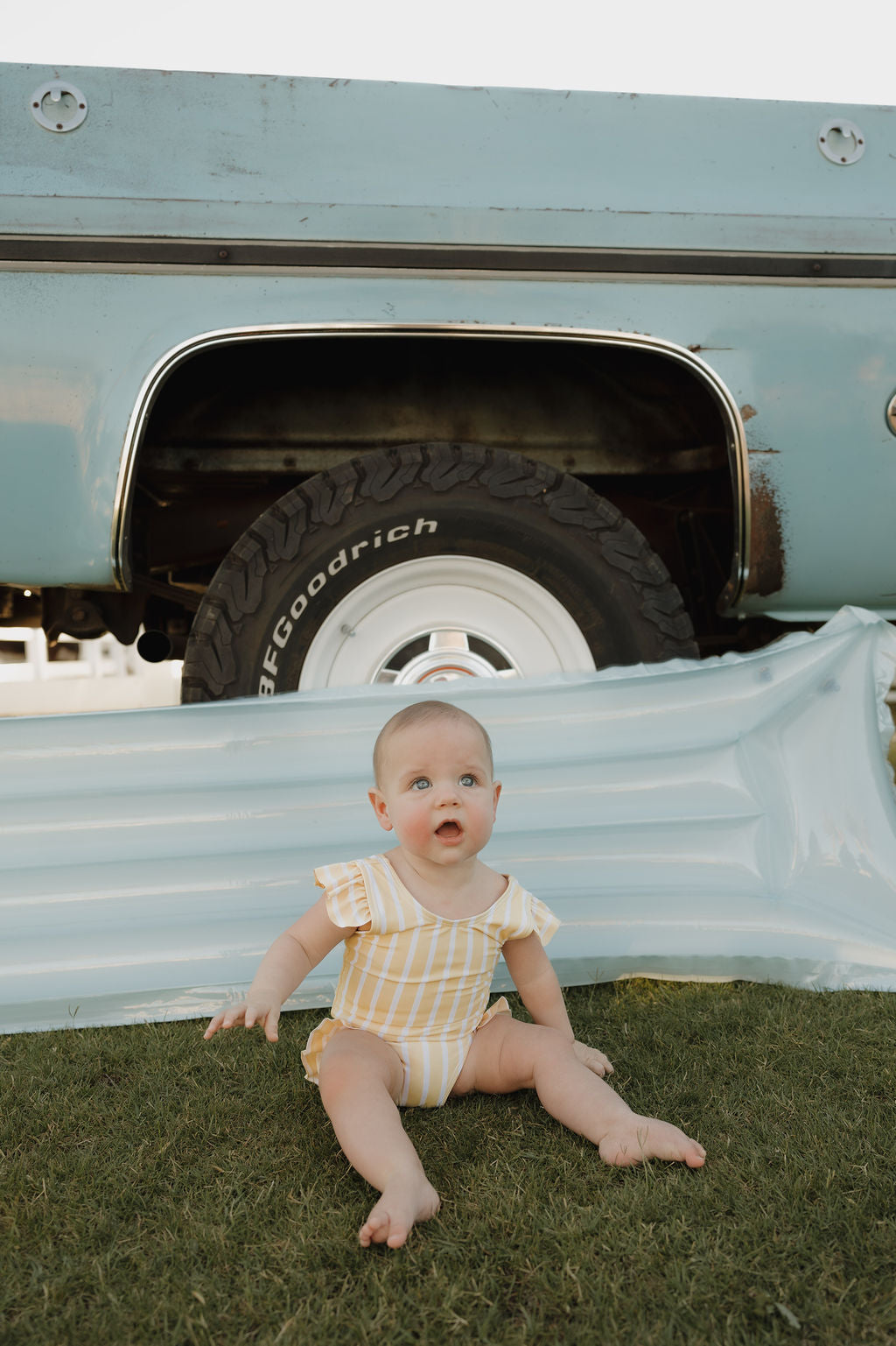 A baby in the lolo webb 2 Girl Sleeveless Swimsuit | Sun Rays, featuring yellow and white stripes with ruffle detail, sits barefoot on grass before a blue inflatable mat and an old blue truck with a visible tire.