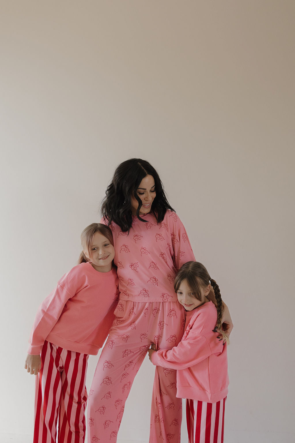 A woman in pink pajamas smiles as two children, dressed in Minty Cherries' Child Sweatshirt | Iconic Cherry with a hand-drawn cherry design, hug her affectionately against a plain background.