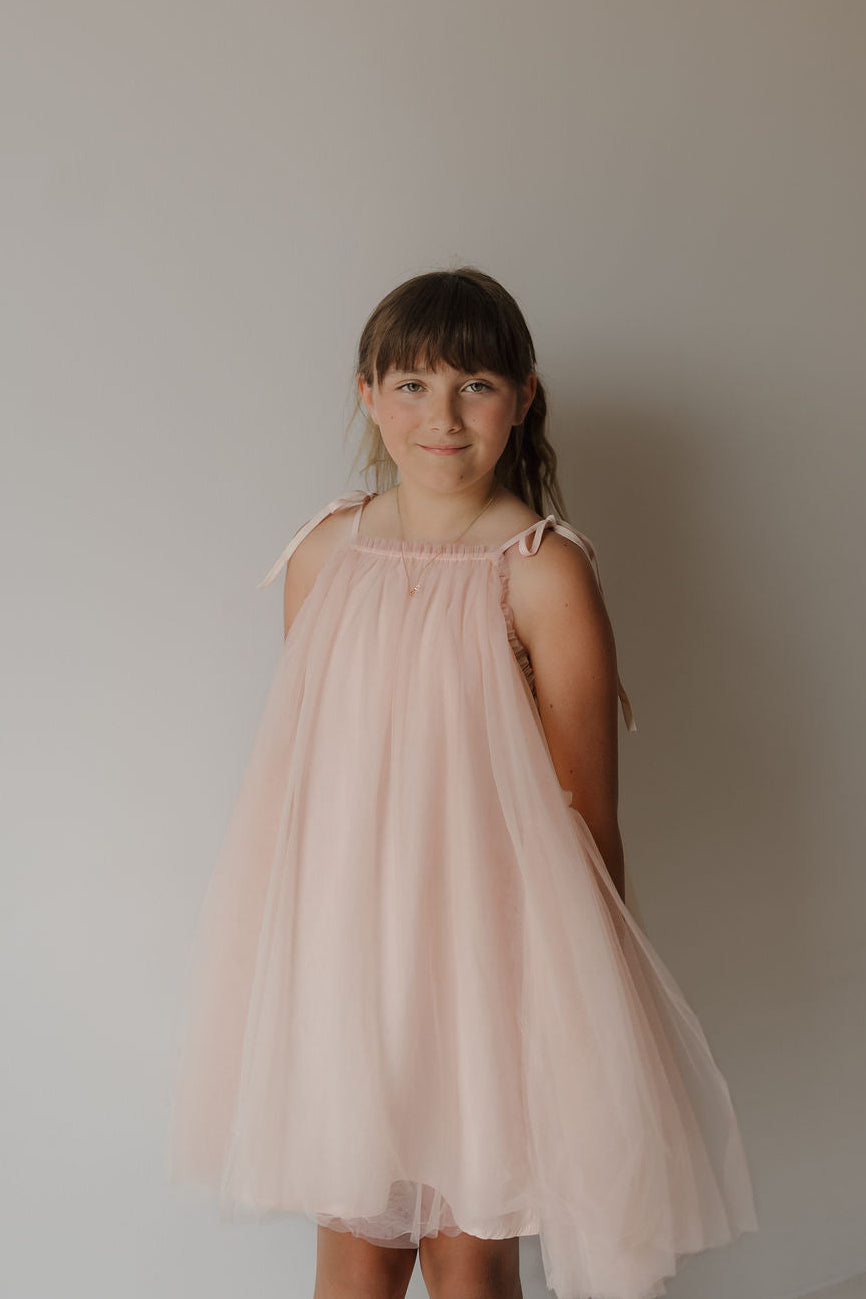 A young girl with brown hair stands against a simple light background, smiling softly at the camera while wearing the Tutu Dress | Slipper by forever french baby— a pale pink, sleeveless tutu dress featuring tied bow straps on her shoulders.