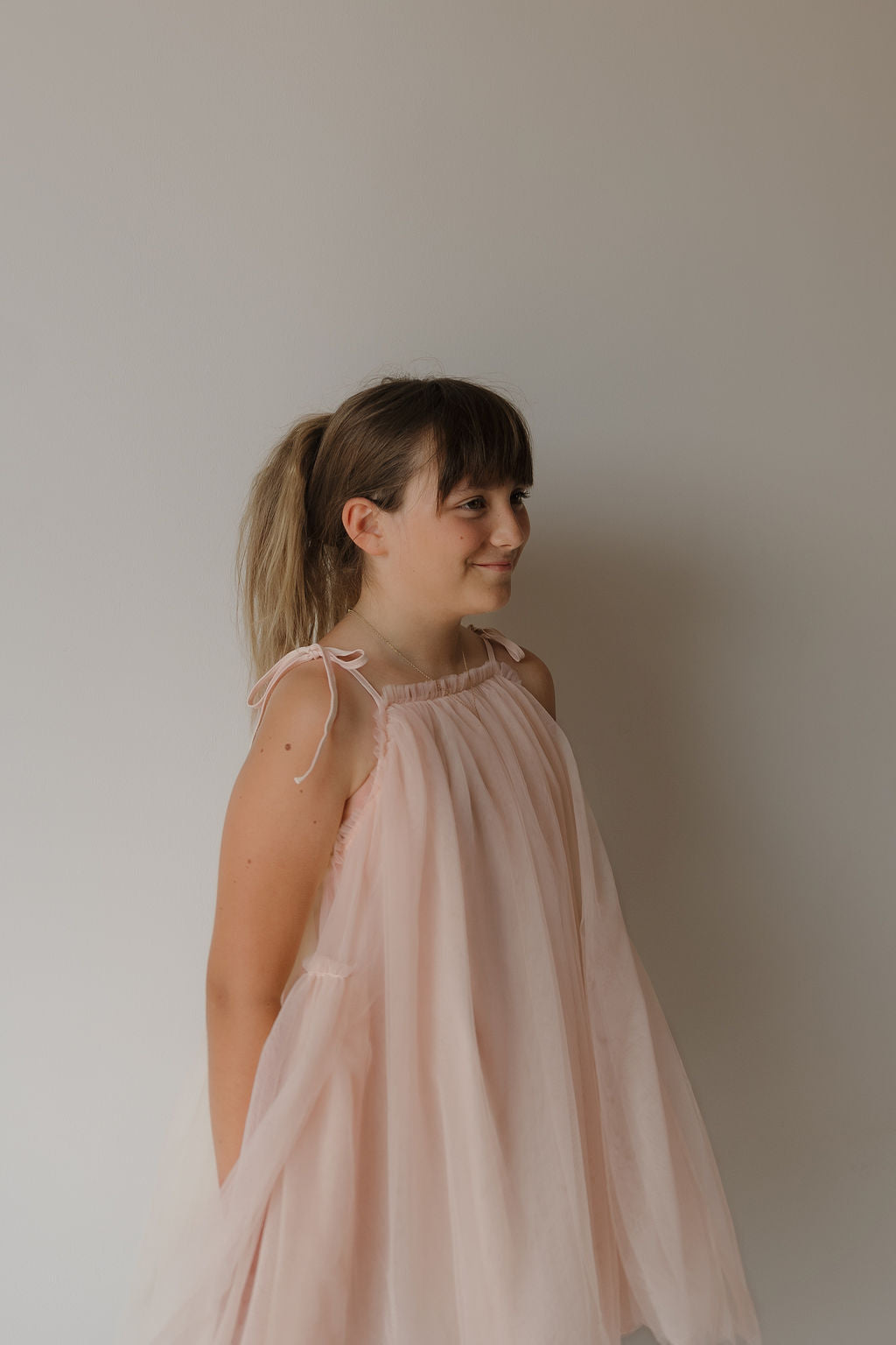 A young girl with light brown hair in a ponytail wears the forever french baby Tutu Dress | Slipper, featuring delicate bow straps, and stands against a plain, light-colored wall, looking to the side with a soft smile.