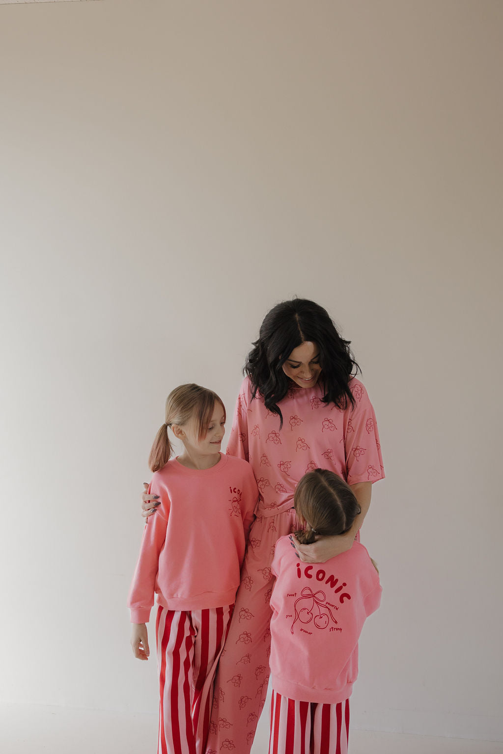 A woman and two young girls smile at each other against a plain background. The girls wear Minty Cherries "Iconic Cherry" sweatshirts featuring a hand-drawn cherry, paired with red-and-white striped pants.