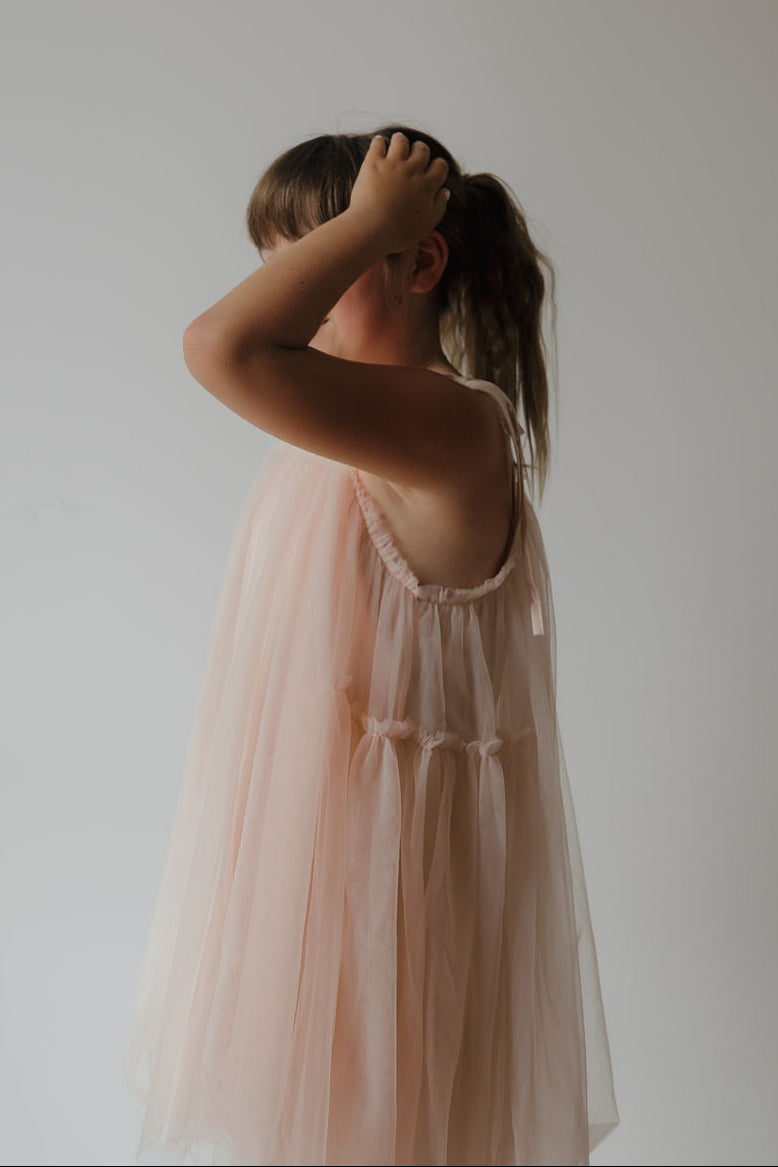 A young girl models the forever french baby Tutu Dress | Slipper in light pink, standing sideways with one hand gently resting on her head, set against a plain, light-colored background.