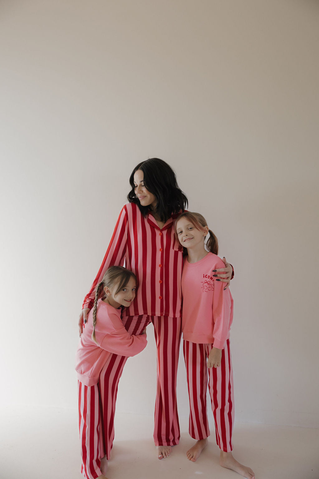 A woman in the Minty Cherries Women's Bamboo Button Up Lounge Set | Iconic Stripe stands barefoot with two girls in matching pajamas, sharing hugs against a plain background—their soft bamboo clothing enhances the cozy moment.