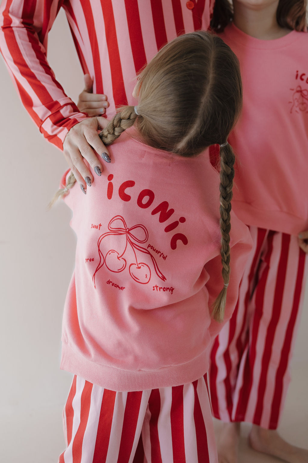Three children wear matching red-and-white striped pajama pants and Minty Cherries “Iconic Cherry” sweatshirts, featuring a hand-drawn cherry design on the back. Two have braided hair; one is partly visible.