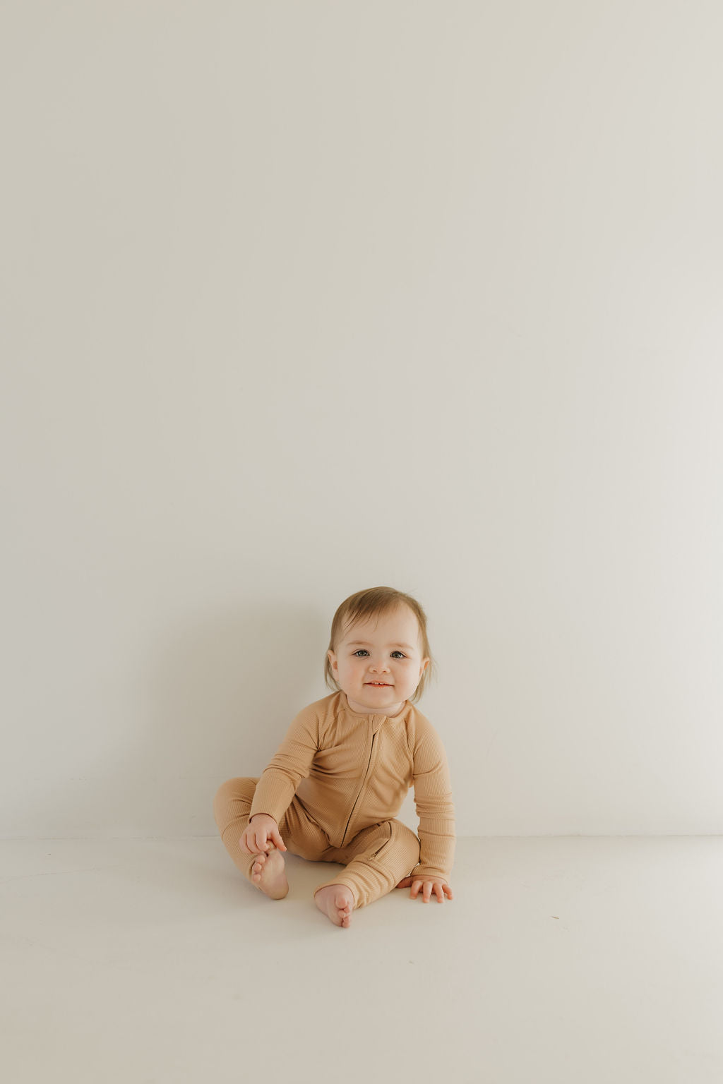 A baby, dressed in forever french baby's Bamboo Zip Pajamas in Tawny, sits contentedly on a light-colored floor against a plain, off-white background, with one hand on the floor and the other gently touching their leg.