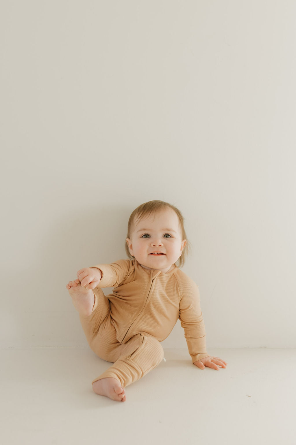 A baby, smiling and playfully holding a foot, sits on a white floor against a white wall while enjoying the comfort of hypo-allergenic Bamboo Zip Pajamas in Tawny by forever french baby.