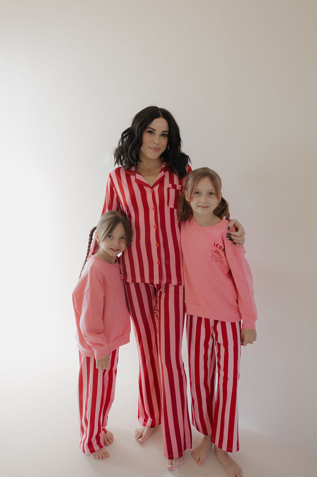 A woman in the Minty Cherries Women's Bamboo Button Up Lounge Set | Iconic Stripe stands barefoot and smiling with two young girls in pink tops and red-and-white striped lounge pants, against a plain light background.