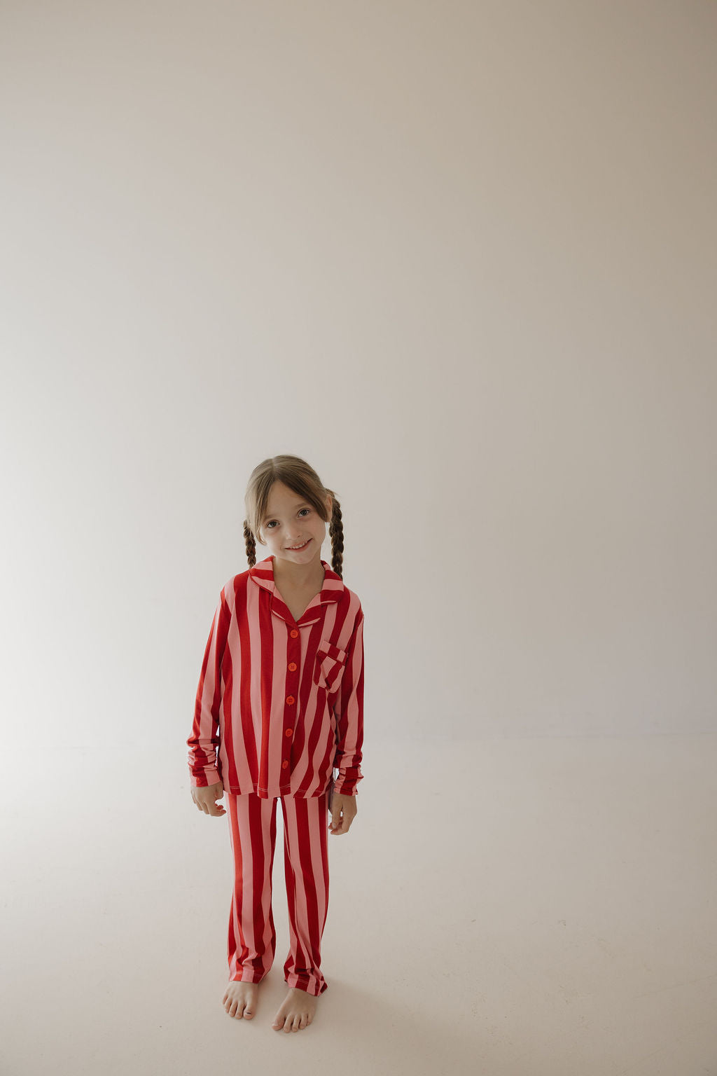 A young child with pigtails stands barefoot against a light backdrop, smiling while wearing the Minty Cherries Toddler Bamboo Button Up Lounge Set in Iconic Stripe—a hypo-allergenic and stylish choice for comfort.