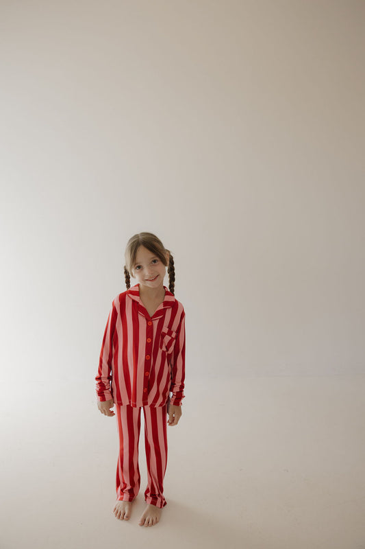 A young child with pigtails stands barefoot against a light backdrop, smiling while wearing the Minty Cherries Toddler Bamboo Button Up Lounge Set in Iconic Stripe—a hypo-allergenic and stylish choice for comfort.