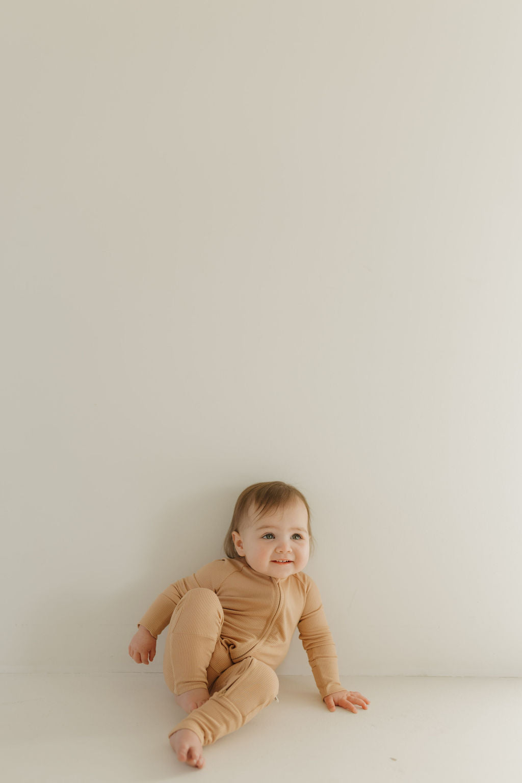 A baby in forever french baby's Bamboo Zip Pajamas | Tawny sits on the floor against a plain white wall, smiling and looking upward. The room's soft lighting accentuates the baby's joyful expression.