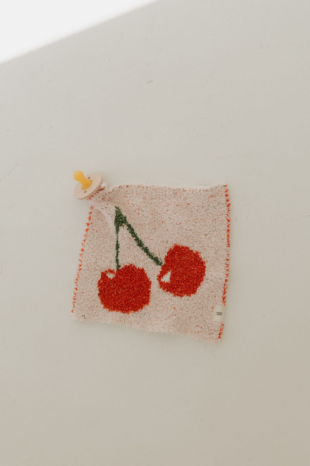 The Lovey | Cherry Orchard, a textured rug with two red cherries on a light background, is pinned to the wall. A beige pacifier from forever french baby rests in the top left corner, with shadows creating soft contrasts around it.