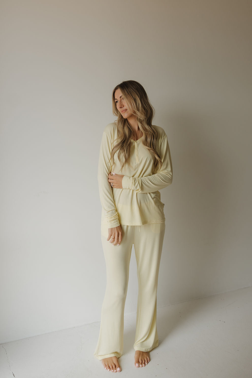 A woman with long wavy hair stands barefoot against a plain white wall, wearing the “Women’s Long Sleeve Bamboo Pajama | Sunny” set from forever french baby—a light yellow ensemble crafted from hypo-allergenic bamboo for a gentle, eco-friendly feel. She gazes to the side, one arm crossed over her body.