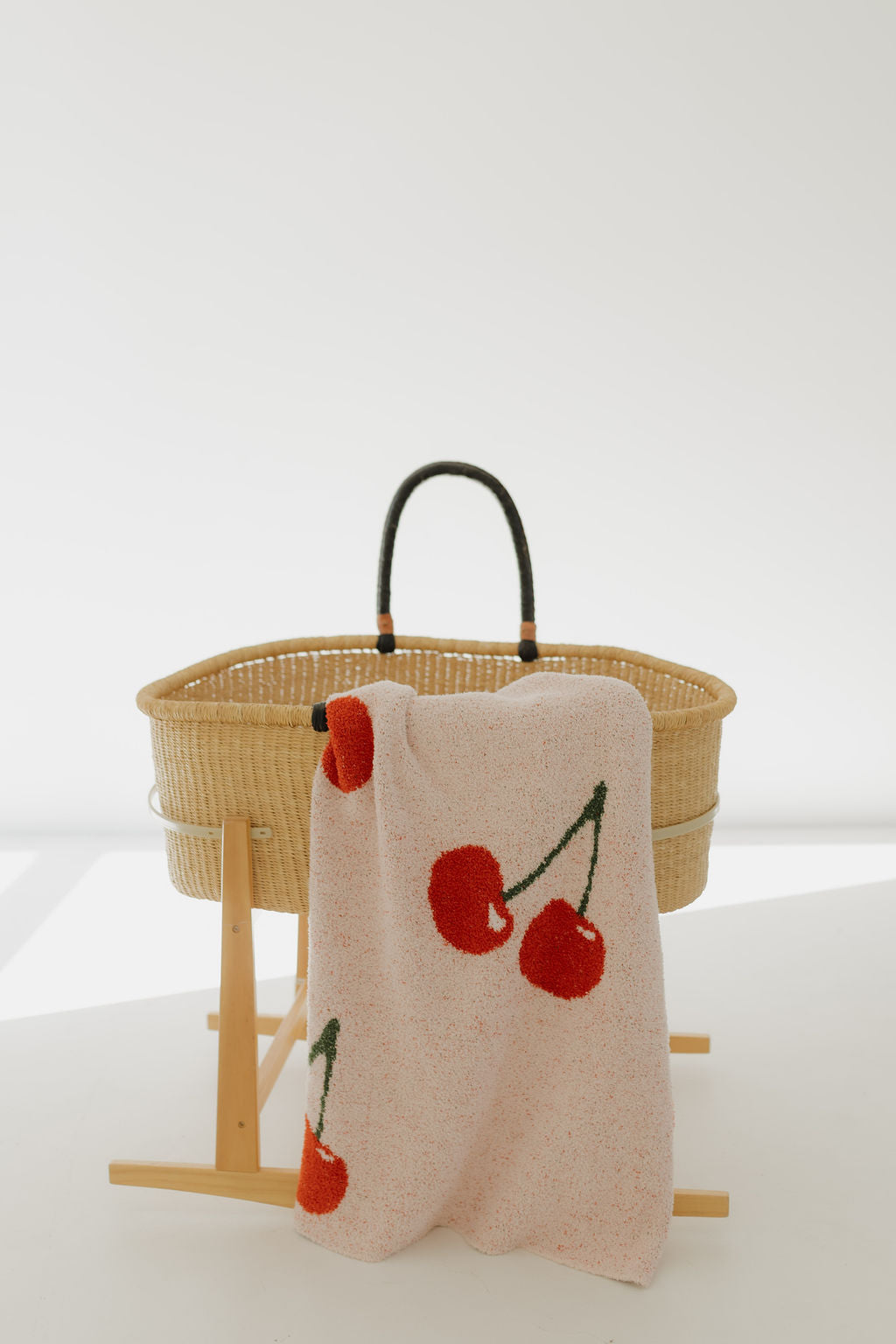 A wicker baby bassinet with black handles rests on a wooden stand. Draped over one side is the Plush Blanket | Cherry Orchard by forever french baby, made from ultra-soft microfiber feather yarn with pink fabric and red cherry designs. The background is bright and minimalistic.