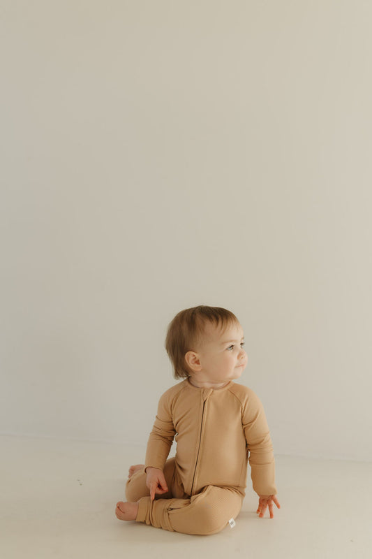 A baby wearing forever french baby's Bamboo Zip Pajamas in Tawny sits on the floor against a plain, light-colored background, gazing calmly to the side.