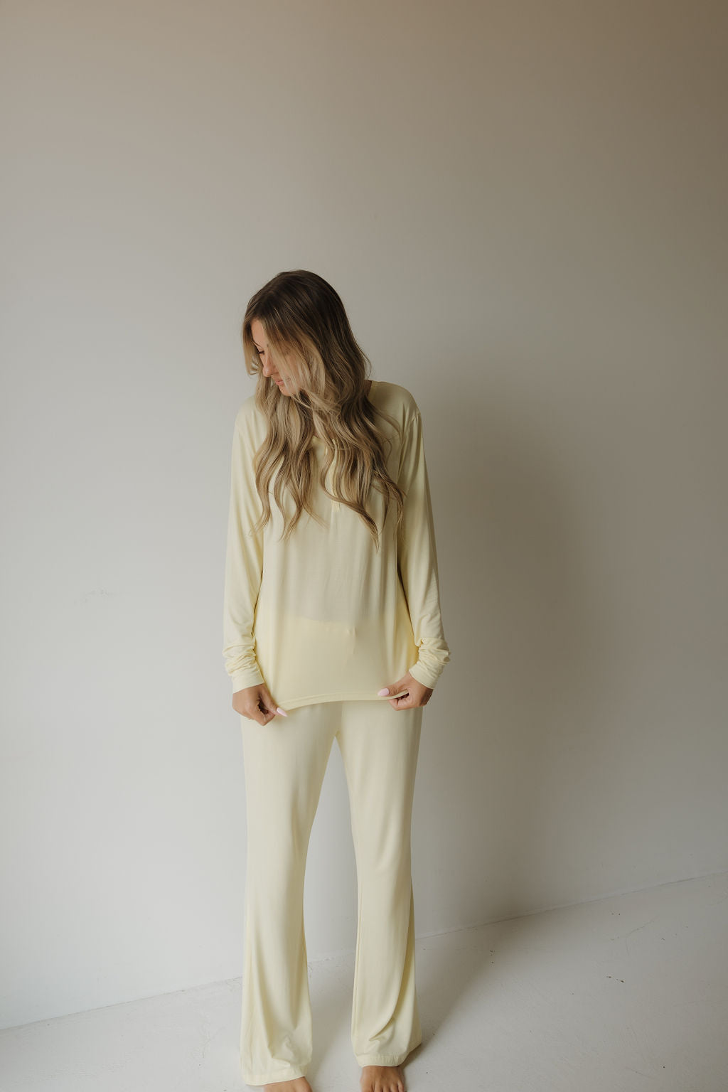 A person with long wavy hair stands barefoot against a plain wall, wearing the forever french baby Women’s Long Sleeve Bamboo Pajama in Sunny—a loose-fitting, light yellow pajama set. This breastfeeding-friendly ensemble features a cozy long-sleeve top and matching pants crafted from hypo-allergenic bamboo fabric, offering both comfort and style.