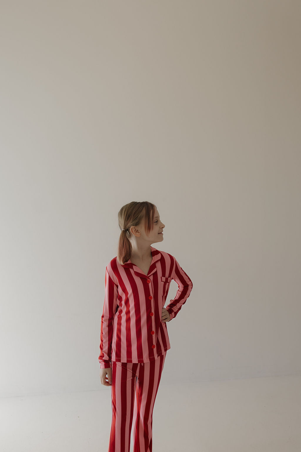 A young child wears the Minty Cherries Toddler Bamboo Button Up Lounge Set in Iconic Stripe, featuring red and white vertical stripes, standing against a plain light wall and looking to the side with one hand on their hip.